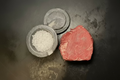 A raw cut of red beef or steak placed on a dark surface, with a small stone bowl containing white salt and a matching stone lid beside it.